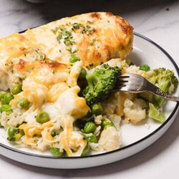 A plate of cheesy baked chicken with rice, peas, broccoli, and cauliflower, with a fork resting on the plate.