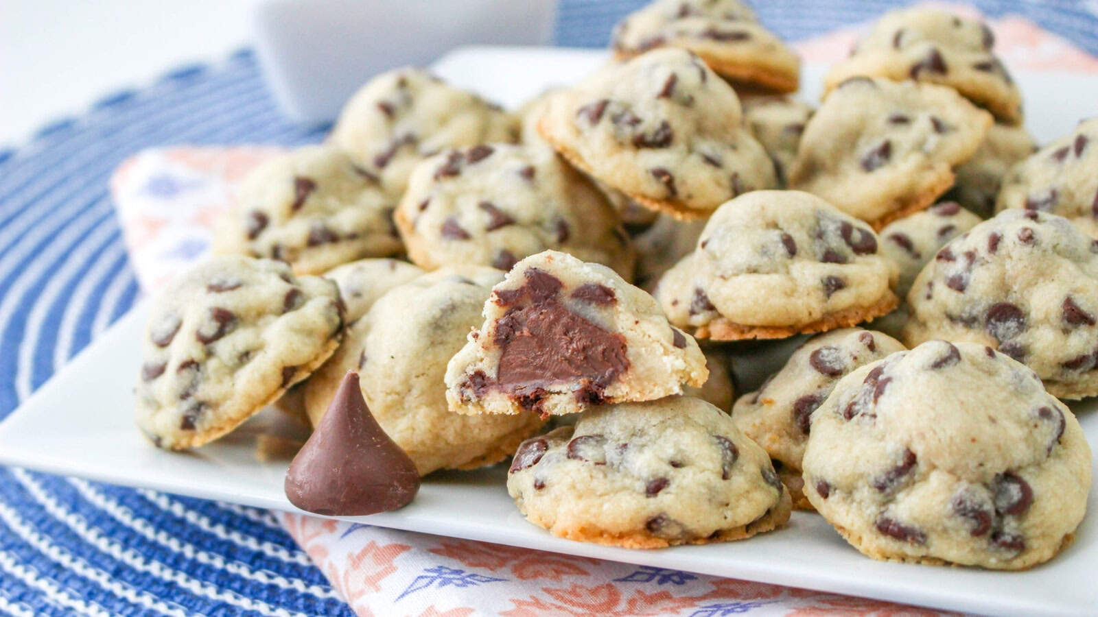 A white plate filled with chocolate chip cookies, some with chocolate filling visible. A chocolate drop is placed in front on a patterned cloth and blue placemat.