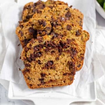 Three slices of zucchini bread with chocolate chips on a white platter, with whole zucchinis and chocolate chips nearby.