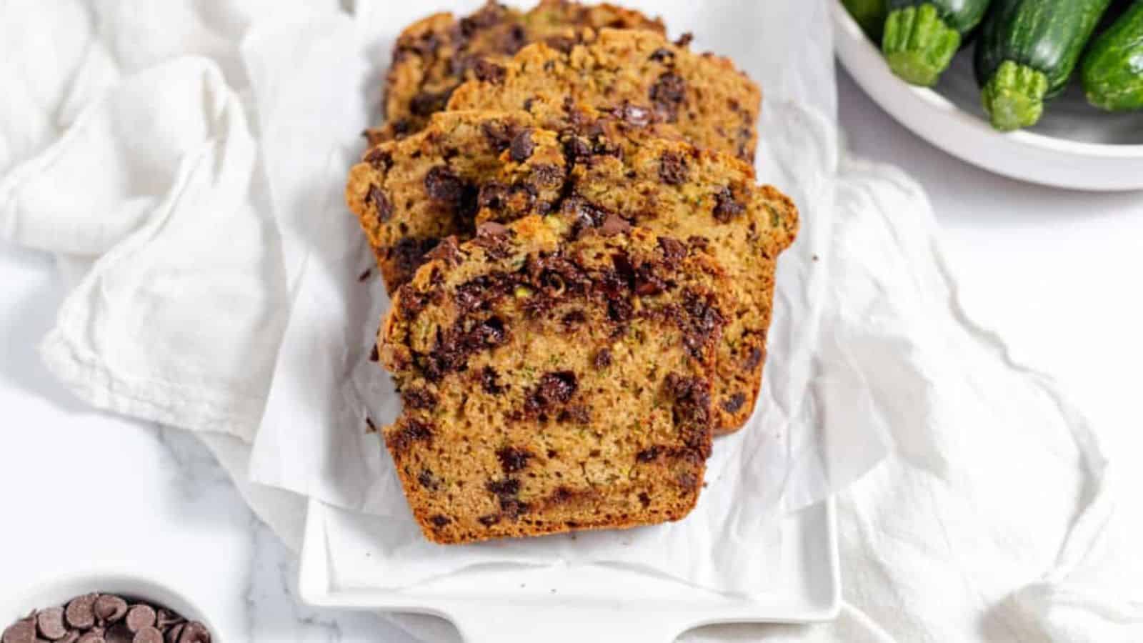 Slices of chocolate chip zucchini bread with chocalate chips and zucchinis in the background.