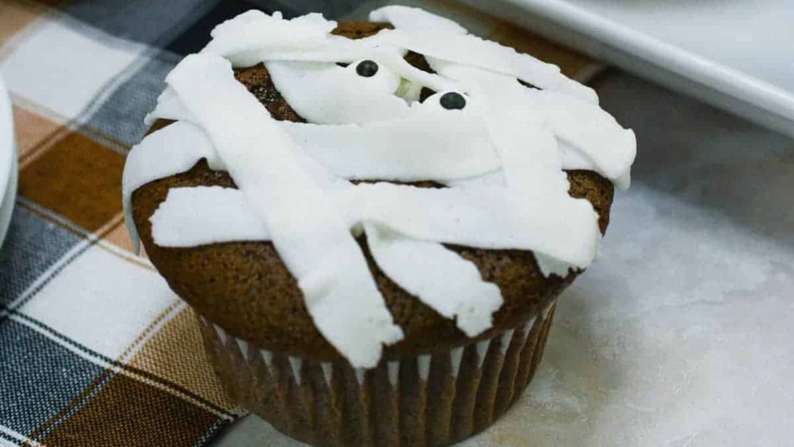 A chocolate mummy cupcake with white frosting on a table.