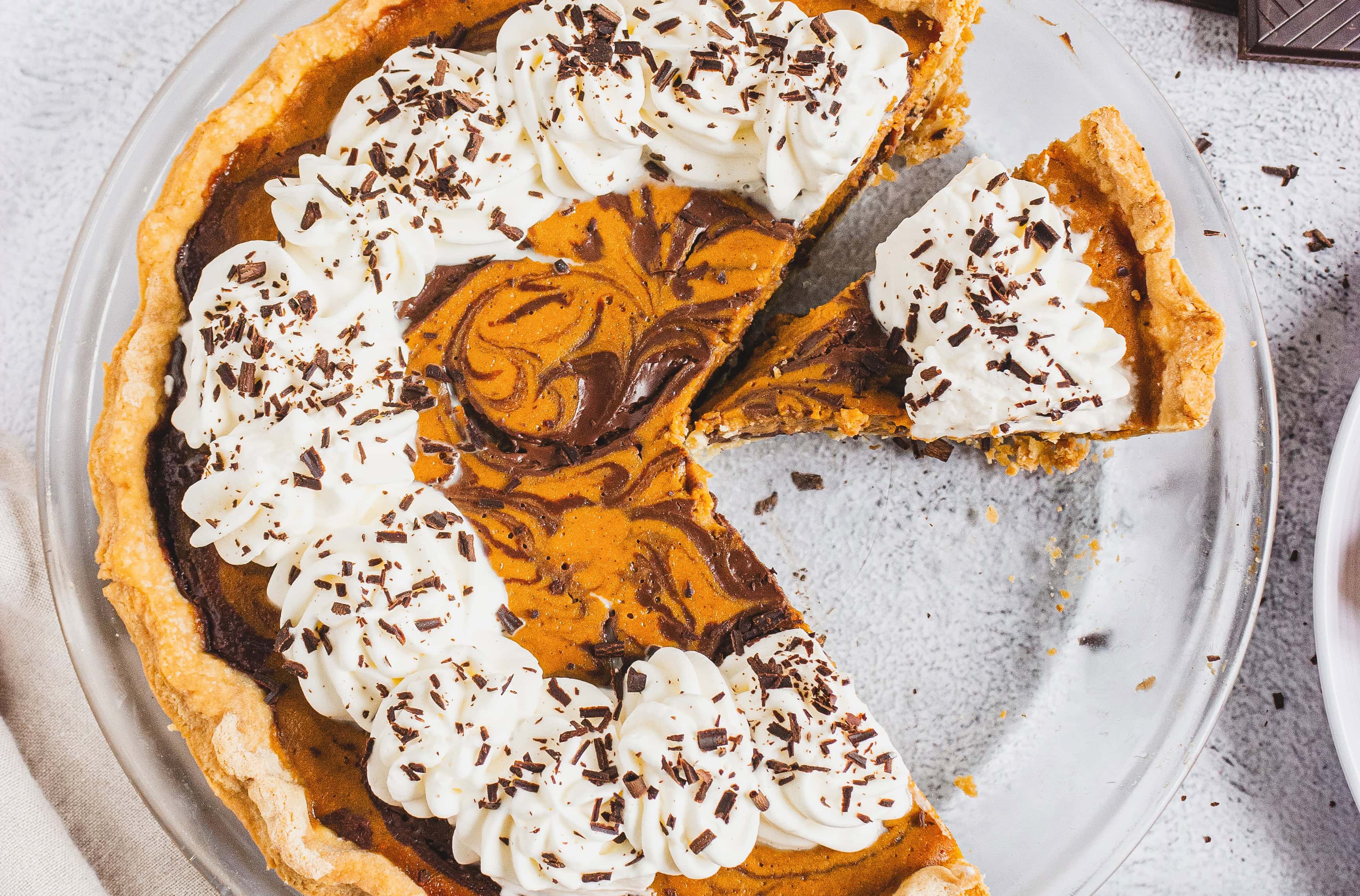 A Chocolate Pumpkin Pie topped with whipped cream and chocolate shavings sits on a glass pie dish, one slice already enjoyed.