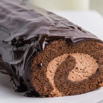Chocolate Swiss roll cake with creamy filling, topped with glossy chocolate ganache, on a white surface.