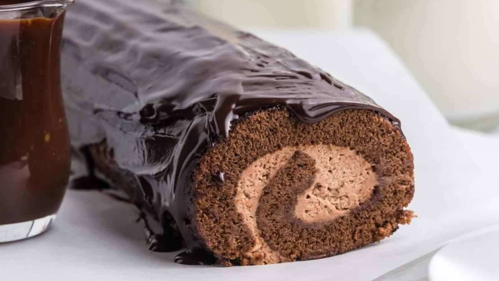 Chocolate swiss roll with three slices cut, revealing cream filling. A small bowl of cocoa and a jug of chocolate sauce are beside it on a white surface.