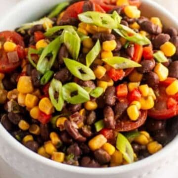 A bowl of black bean salad with corn, cherry tomatoes, and sliced green onions, served with a spoon.
