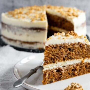 A slice of carrot cake with cream cheese frosting on a plate, garnished with walnuts. The whole cake is in the background.
