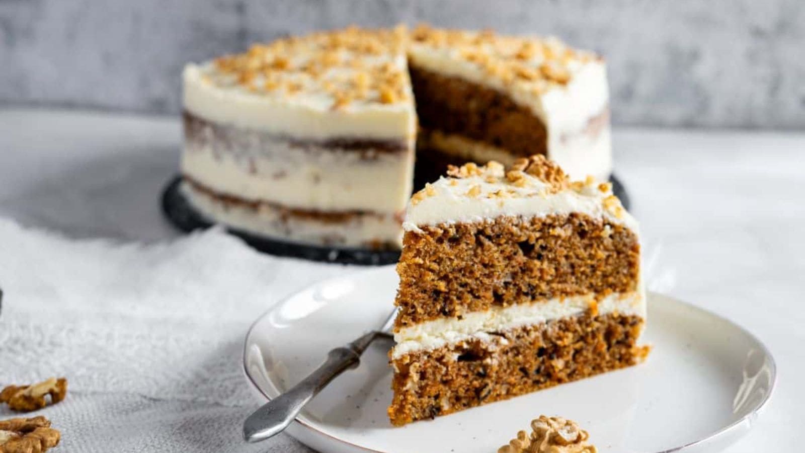 A slice of carrot cake with cream cheese frosting on a plate, garnished with walnuts. The whole cake is in the background.