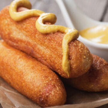 Three corn dogs on paper, one topped with mustard, with a bowl of mustard in the background—amid recent news of a corn dogs recall due to possible contamination.
