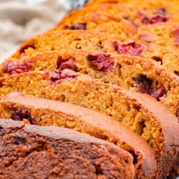 Sliced cranberry bread loaf on a white plate, showing its golden brown crust and berry pieces inside.