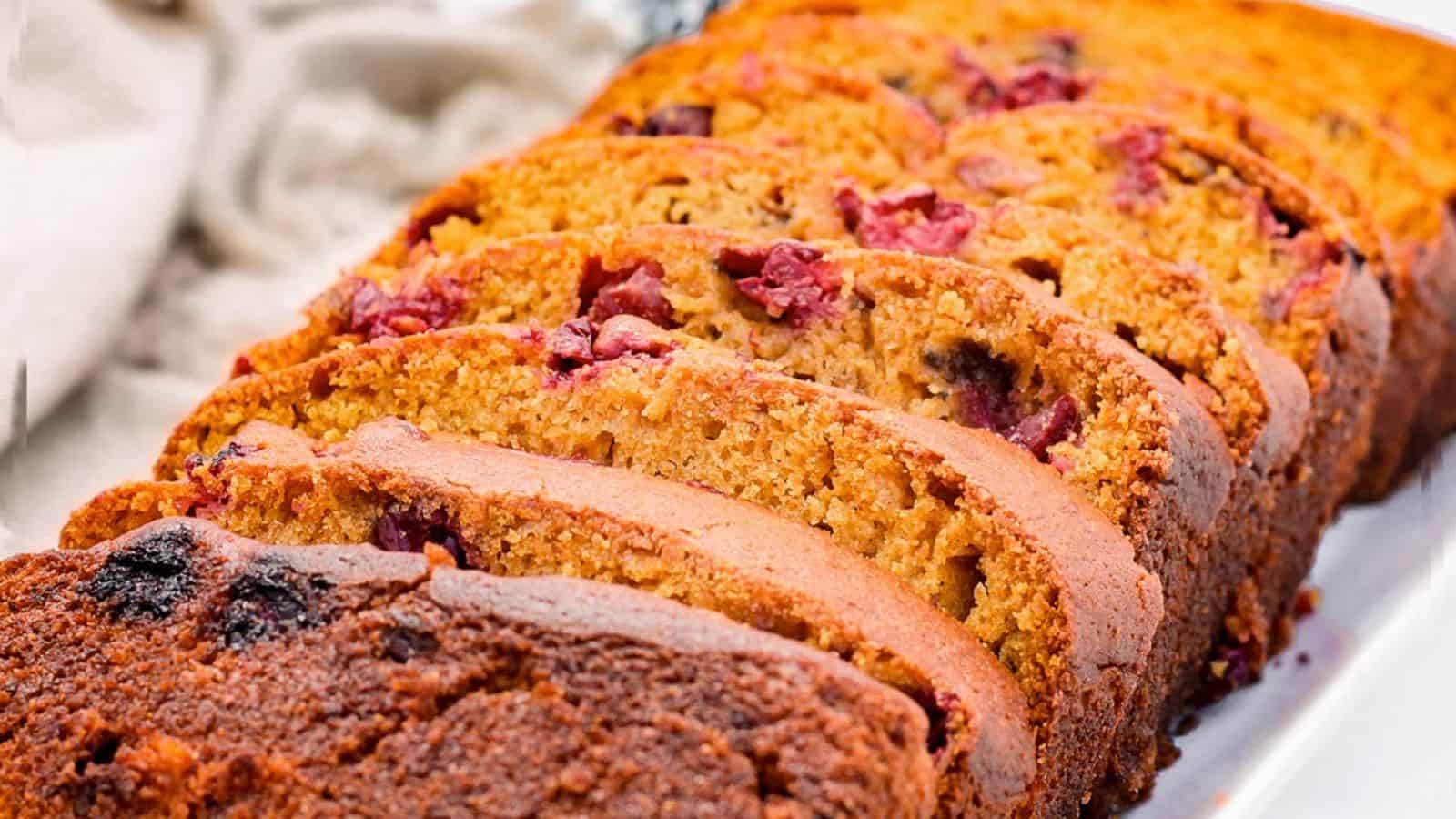 Slices of pumpkin cranberry banana bread arranged on a white plate showcase their golden-brown crust and speckled red berries, creating a warm, inviting scene.