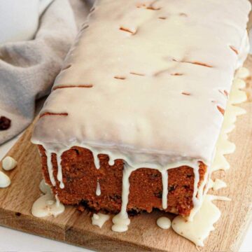 A loaf cake with white icing drips on a wooden board, next to two forks and some dried fruit pieces.