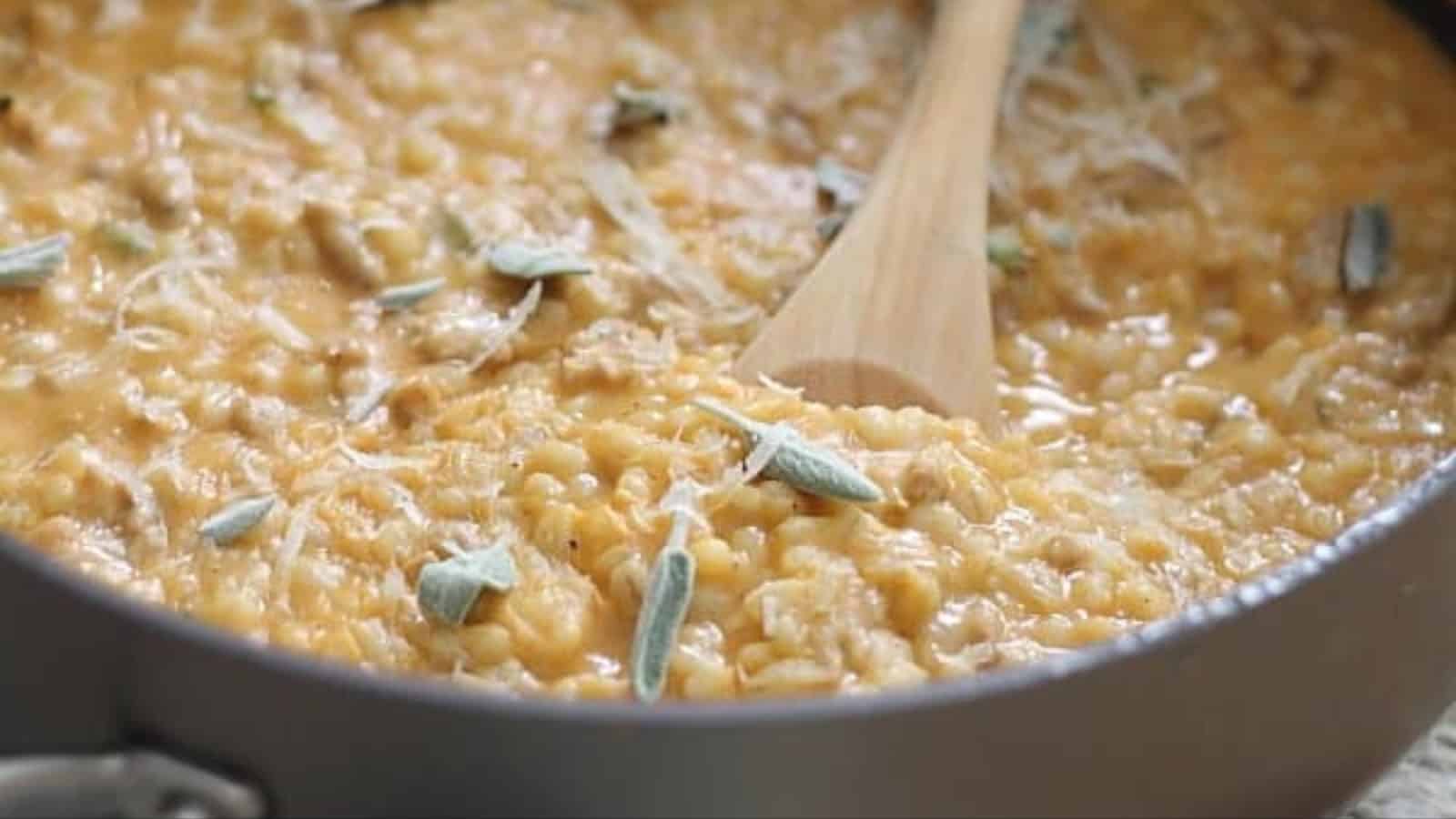 Creamy pumpkin barley with sausage garnished with fresh sage leaves and grated parmesan in a bowl with a spoon.
