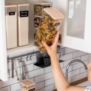 Person placing a clear container of colorful pasta into a kitchen cabinet with labeled food storage containers.