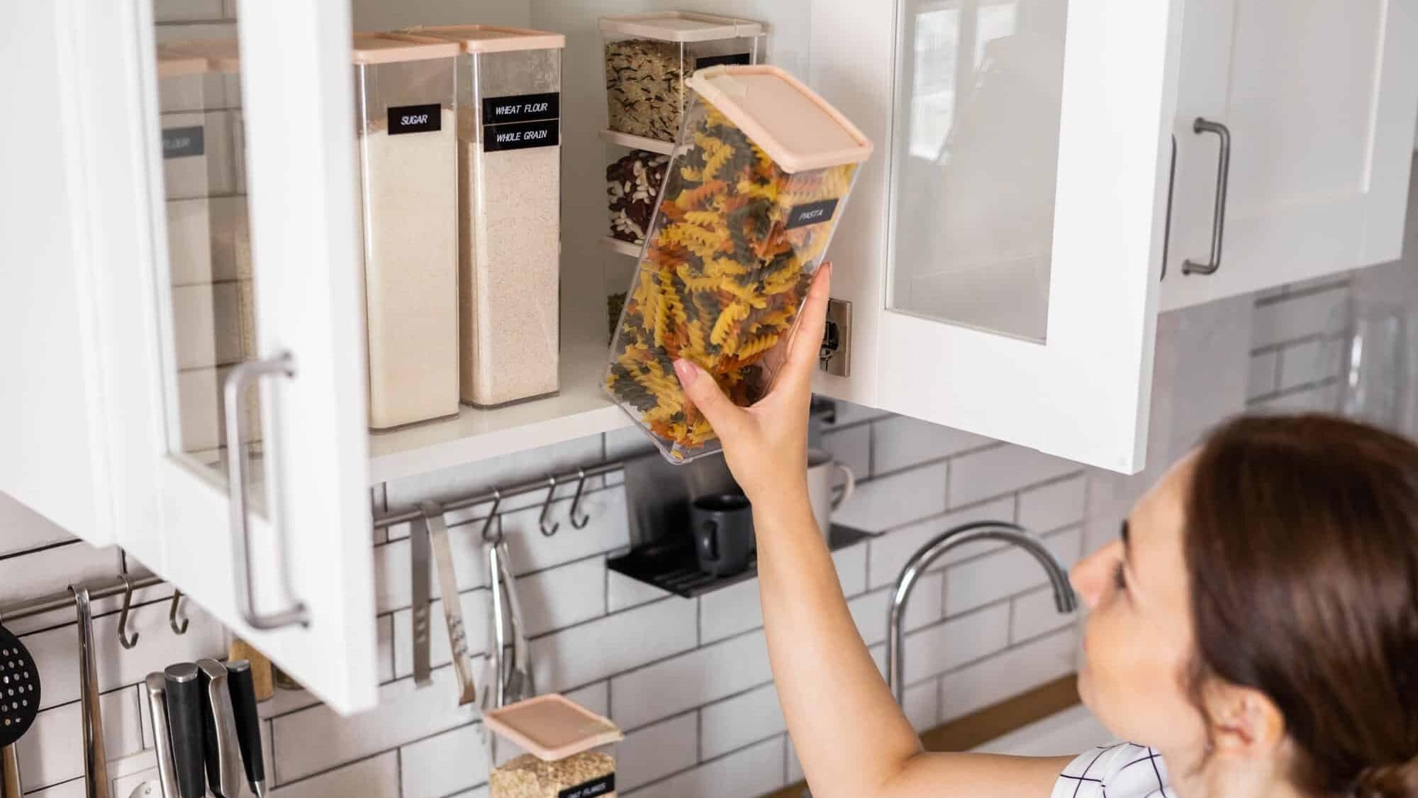 Person placing a clear container of colorful pasta into a kitchen cabinet with labeled food storage containers.