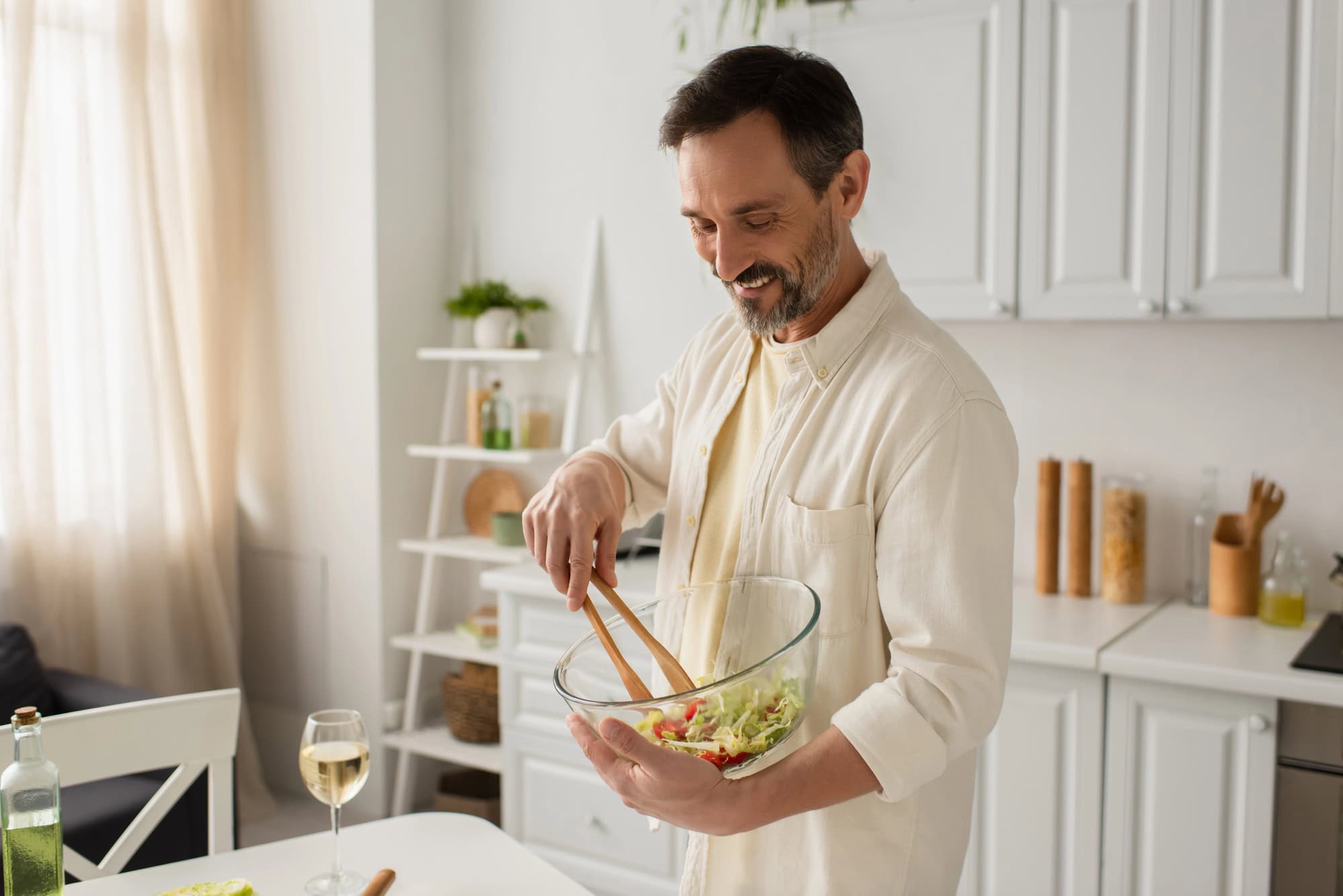 Man smiling while mixing salad in a glass bowl with essential cooking tools for beginners in a bright, modern kitchen and a glass of wine nearby.