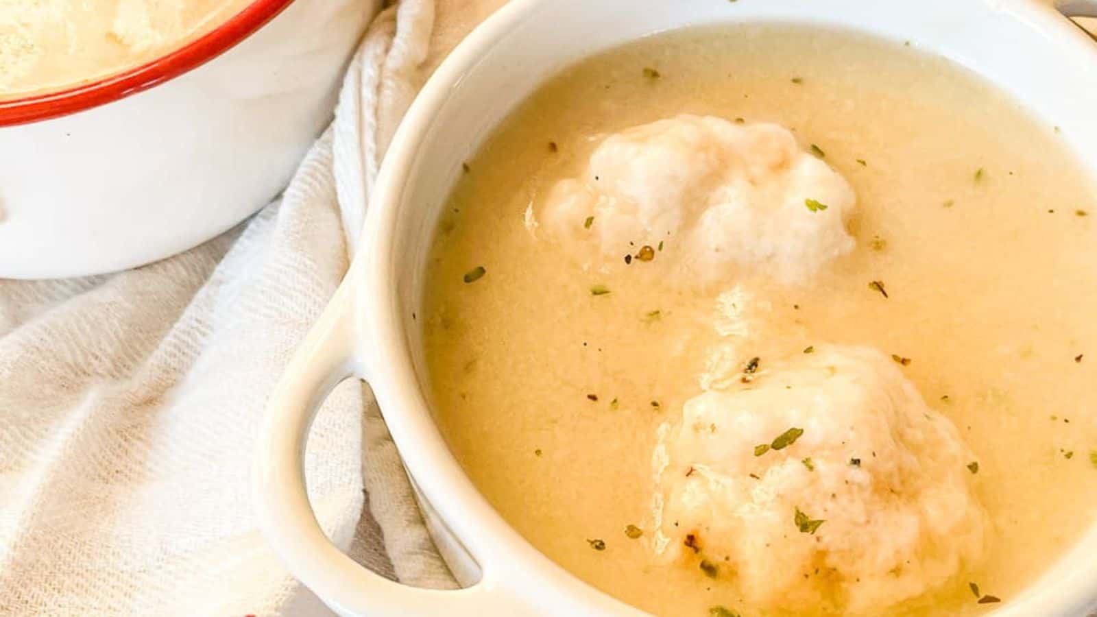 White bowl of creamy soup with two fluffy dumplings, sprinkled with herbs, on a white towel.