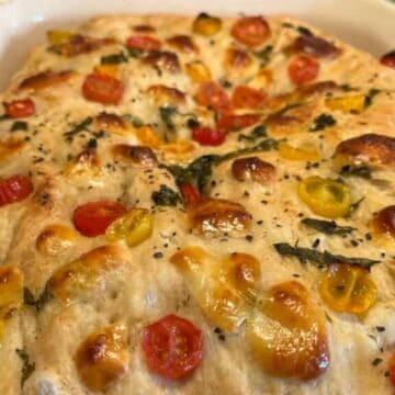 Golden baked focaccia topped with red and yellow cherry tomatoes and fresh herbs in a white baking dish.