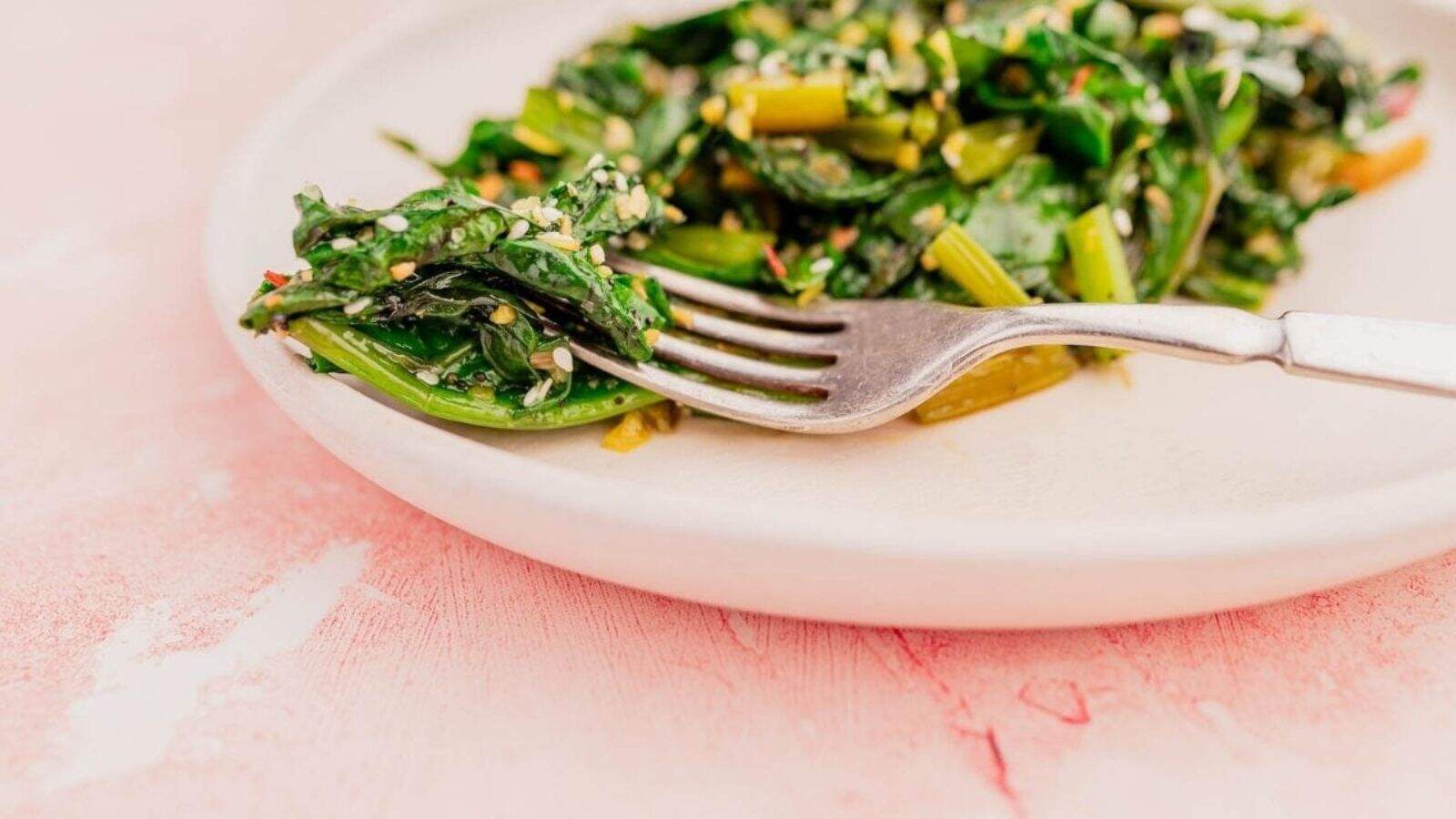 A fork rests on a plate of saut&eacute;ed greens with sesame seeds on a pink surface.