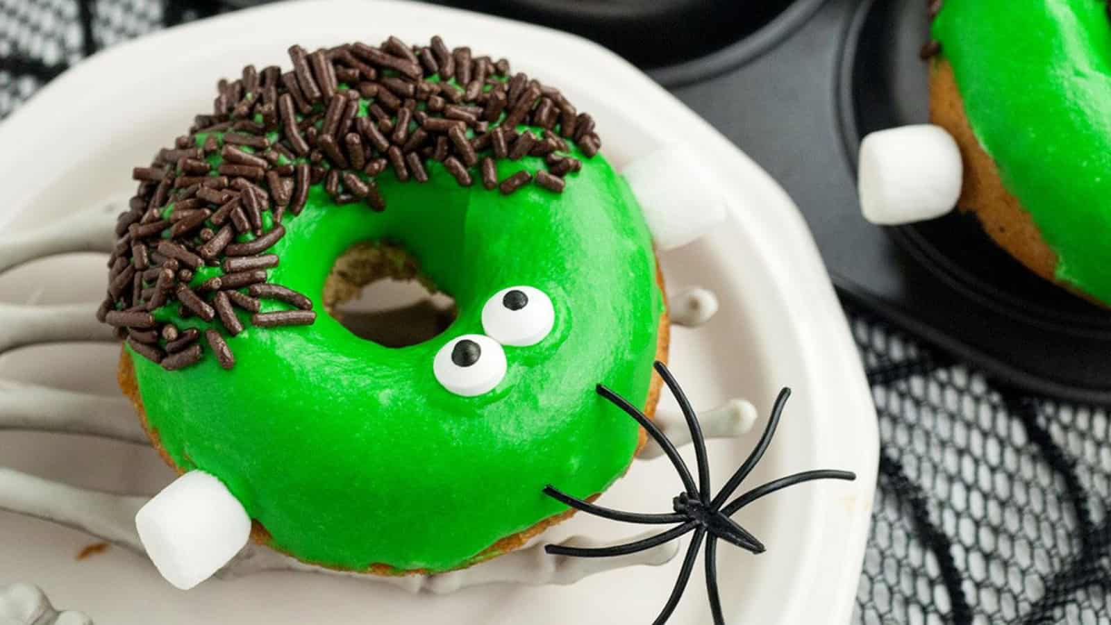 Green Frankenstein-themed donuts with candy eyes, chocolate sprinkles, and marshmallow &ldquo;bolts&rdquo; on a plate.