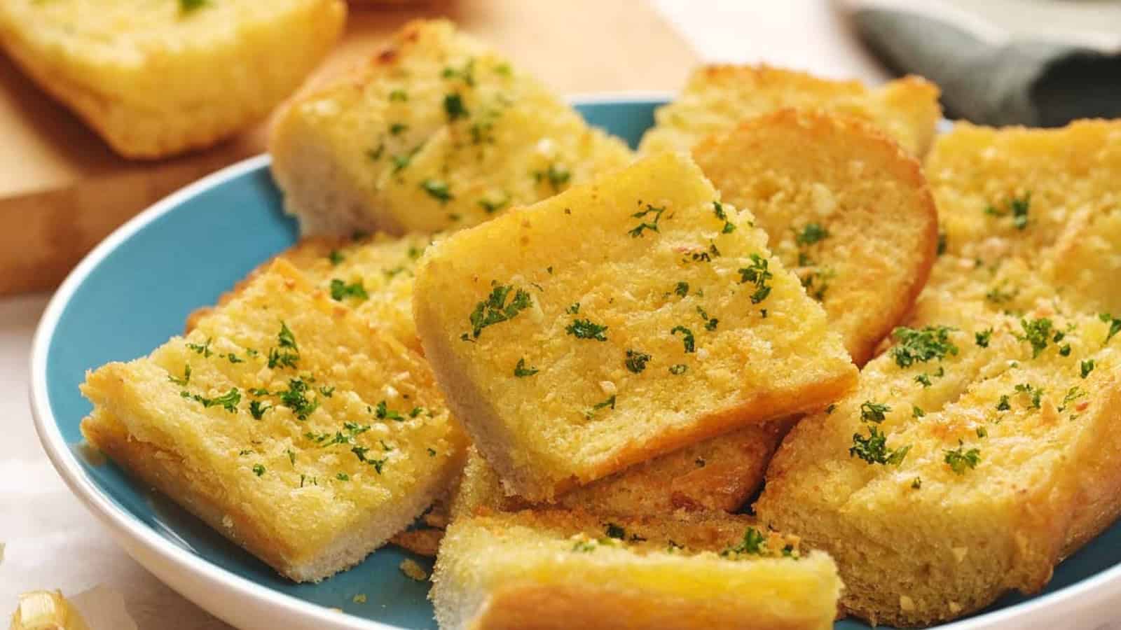 Slices of garlic bread topped with parsley on a blue plate, surrounded by garlic cloves.