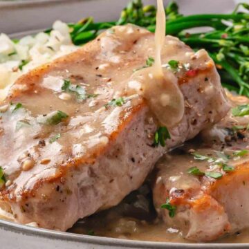 Two pork chops topped with creamy gravy, served with rice and steamed broccolini on a plate.