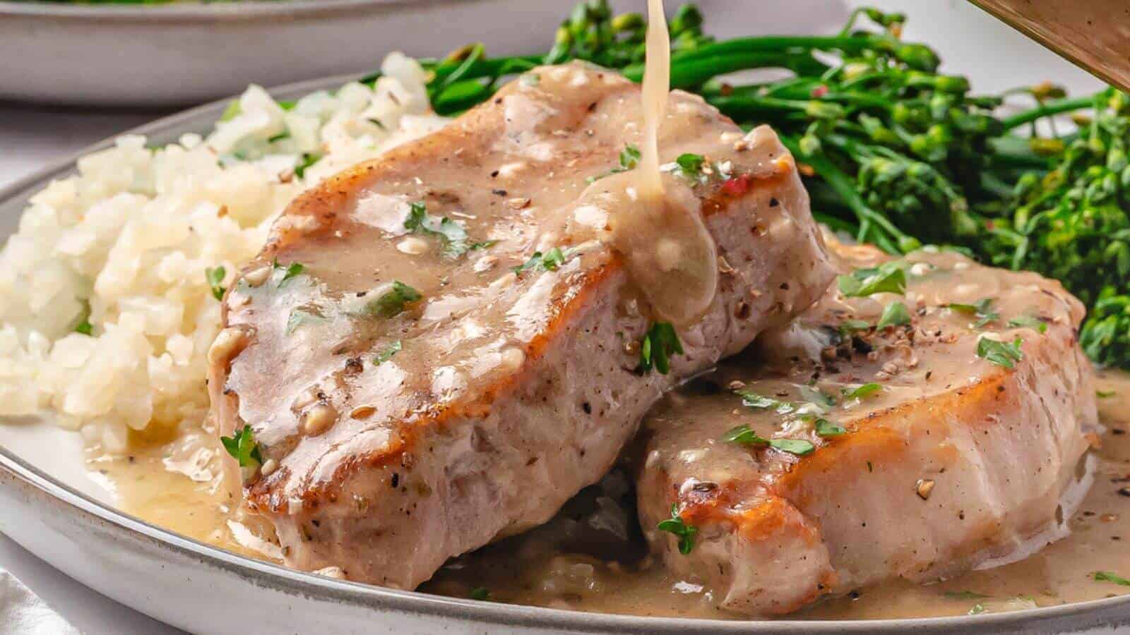 Two pork chops topped with creamy gravy, served with rice and steamed broccolini on a plate.