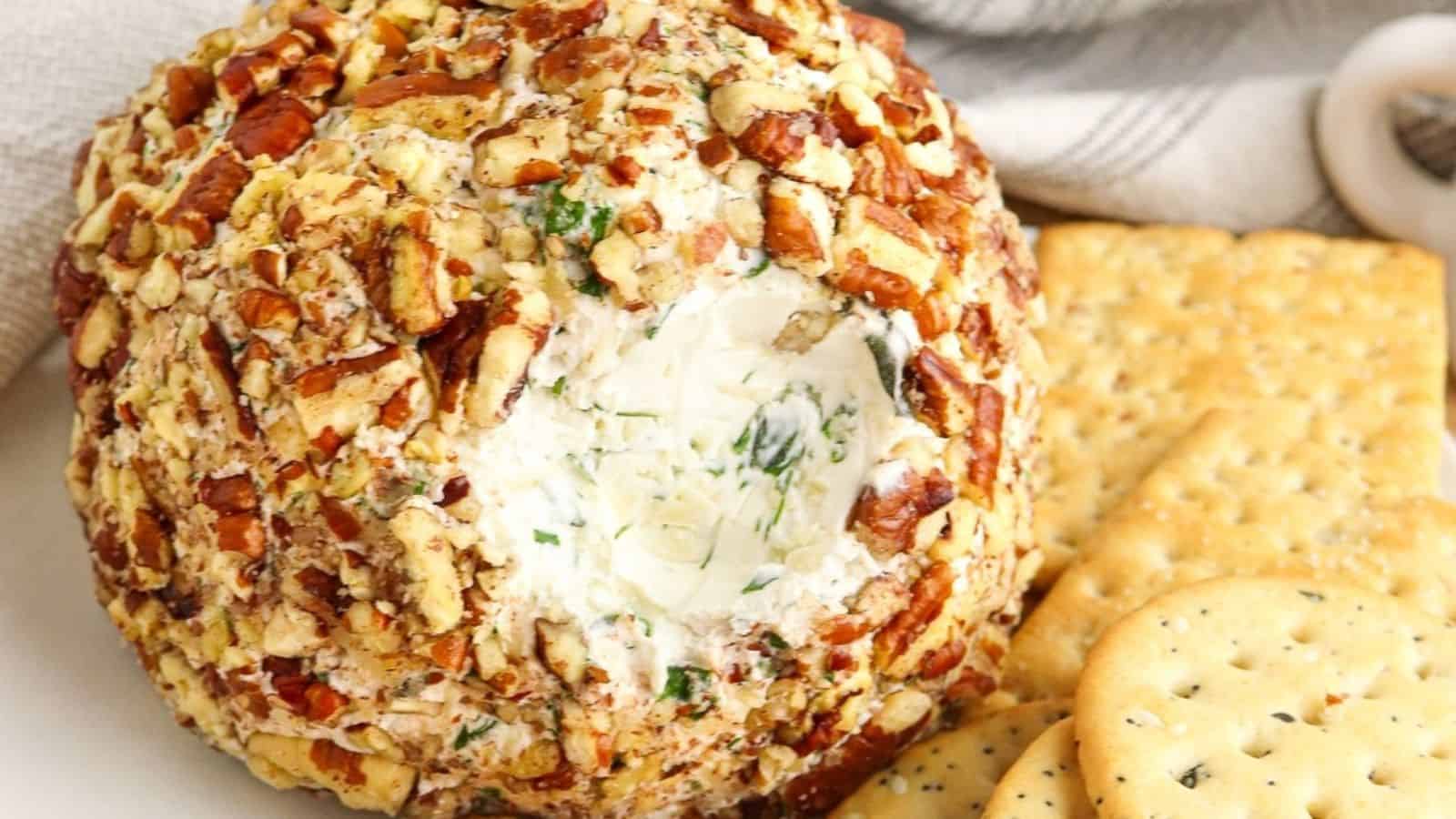 A cheese ball coated in chopped nuts, served with round and rectangular crackers on a white plate.