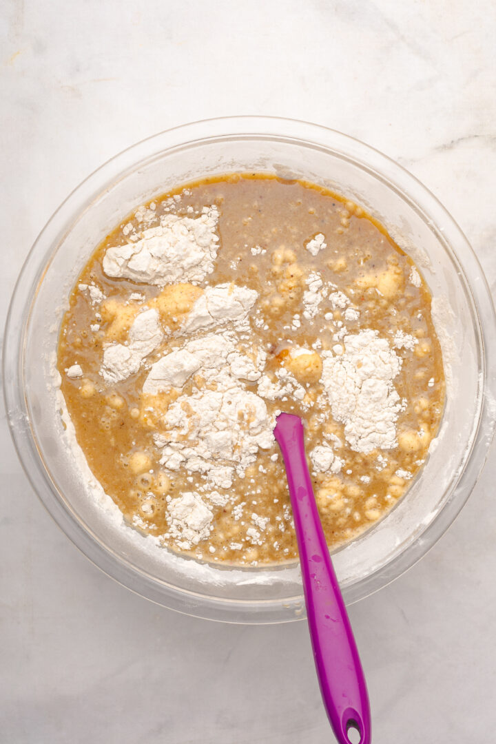 Clear glass bowl with flour, wet ingredients, and a purple spatula on a white surface, ready to mix for a delicious Gingerbread Loaf.