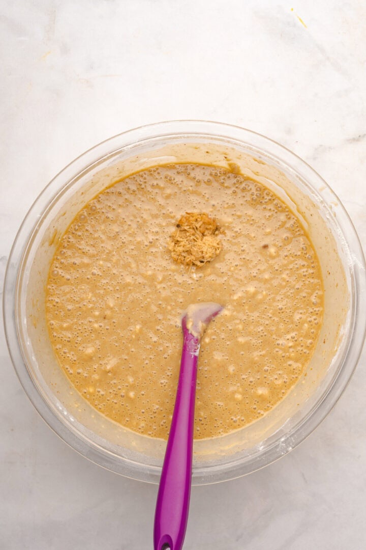 A glass bowl of bubbly gingerbread loaf batter with a purple spatula rests on a white surface.