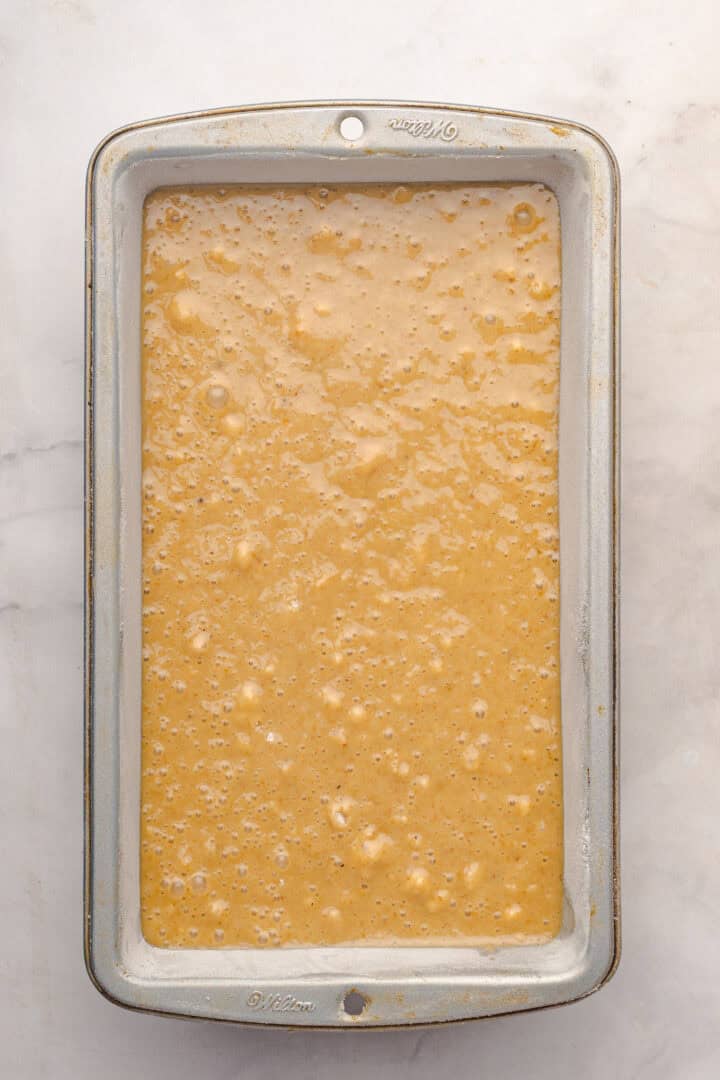 A metal baking pan filled with unbaked, bubbly yellow cake batter—perfect for a classic recipe or even a twist like Gingerbread Loaf—rests on a light surface.