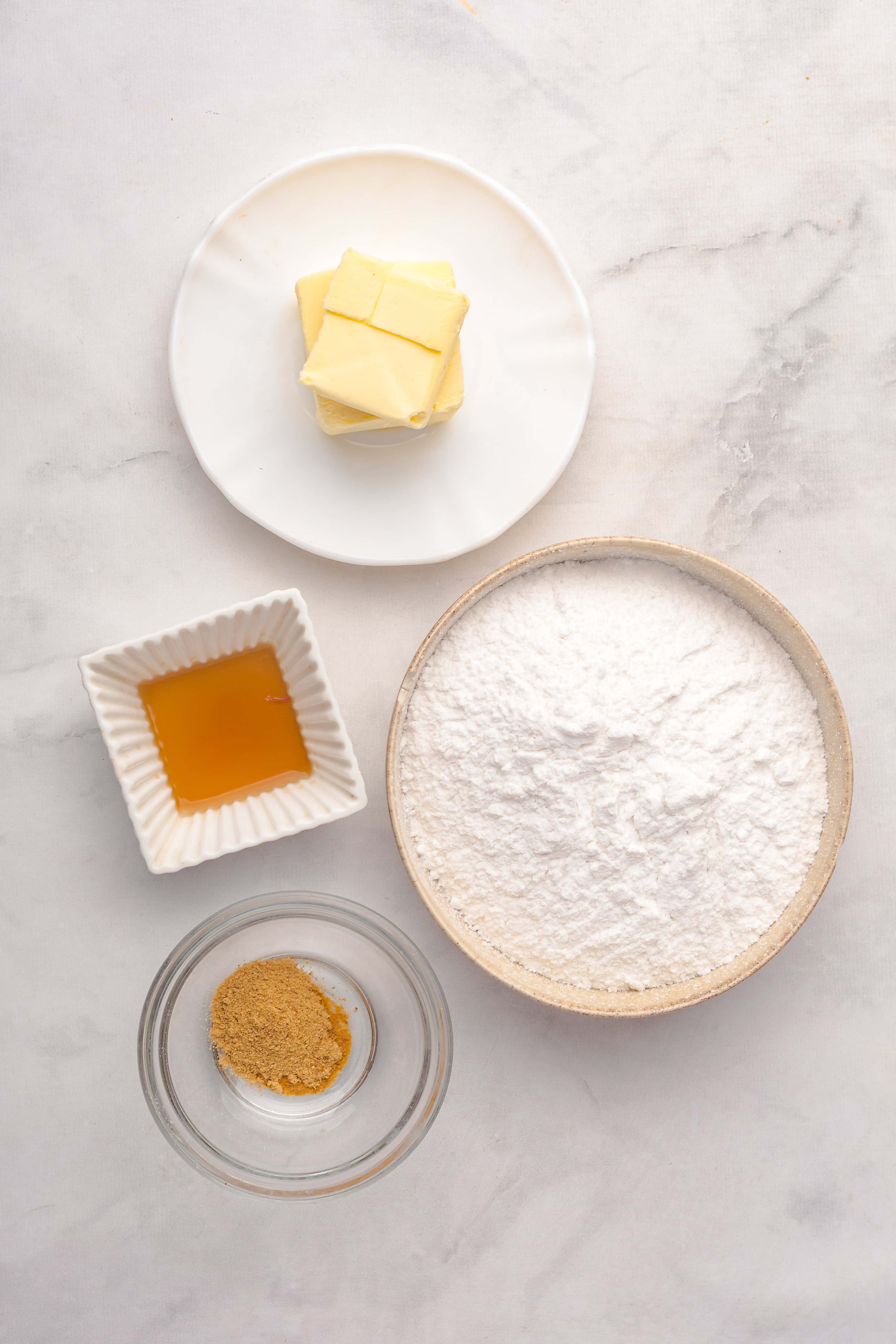 Four bowls with butter, powdered sugar, brown sugar, and vanilla extract on a white surface—perfect ingredients for preparing a delicious Gingerbread Loaf.