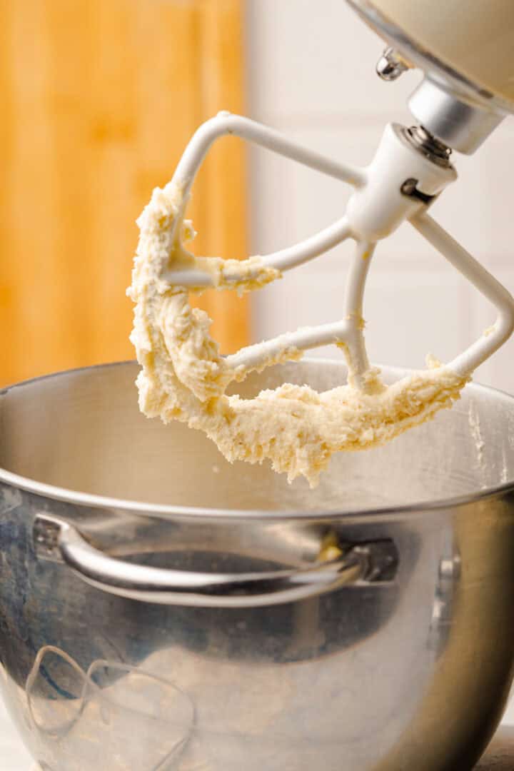A stand mixer paddle with Gingerbread Loaf cookie dough hangs above a stainless steel mixing bowl in a cozy kitchen.