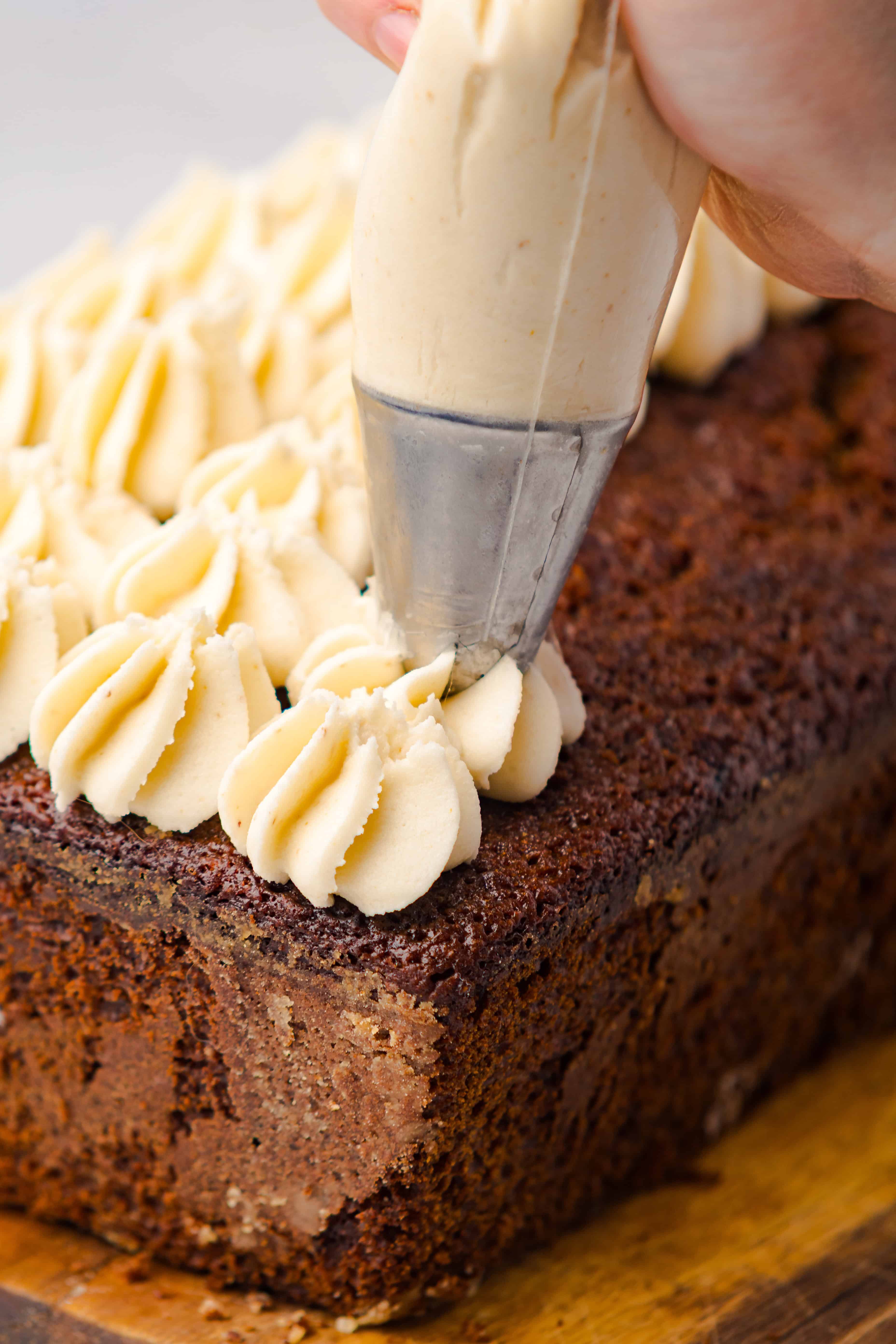A hand pipes swirls of frosting onto a rectangular chocolate cake, resembling a festive Gingerbread Loaf, on a wooden board.