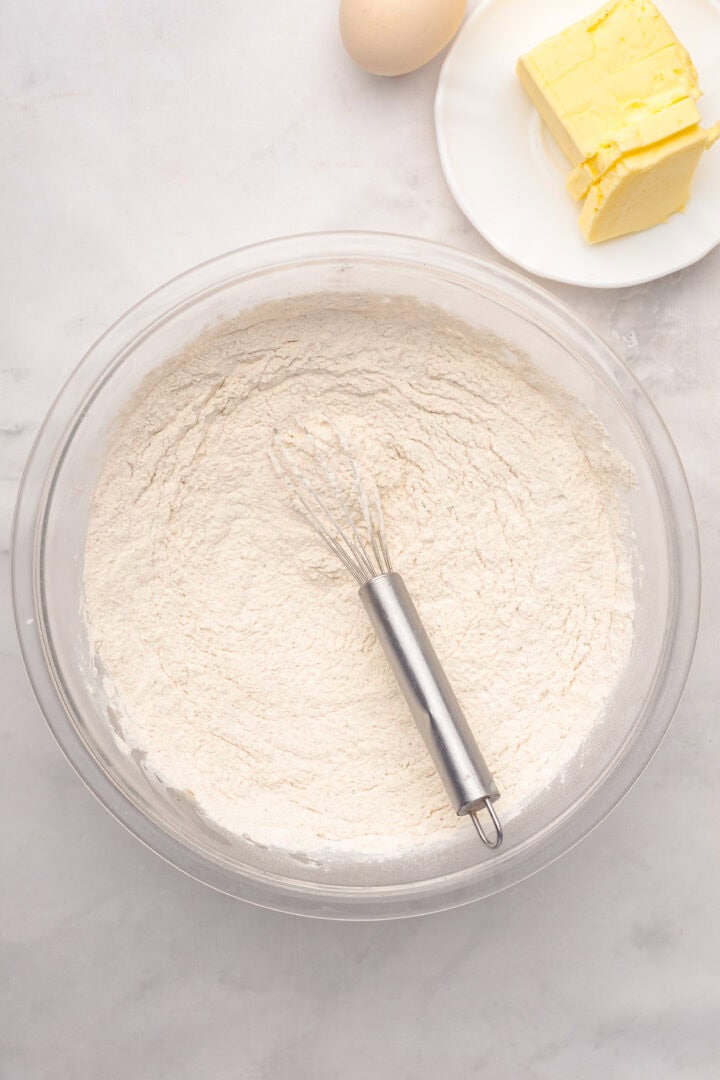 A whisk rests in a bowl of flour as butter and an egg wait on a plate nearby, ready to be transformed into a delicious Gingerbread Loaf.