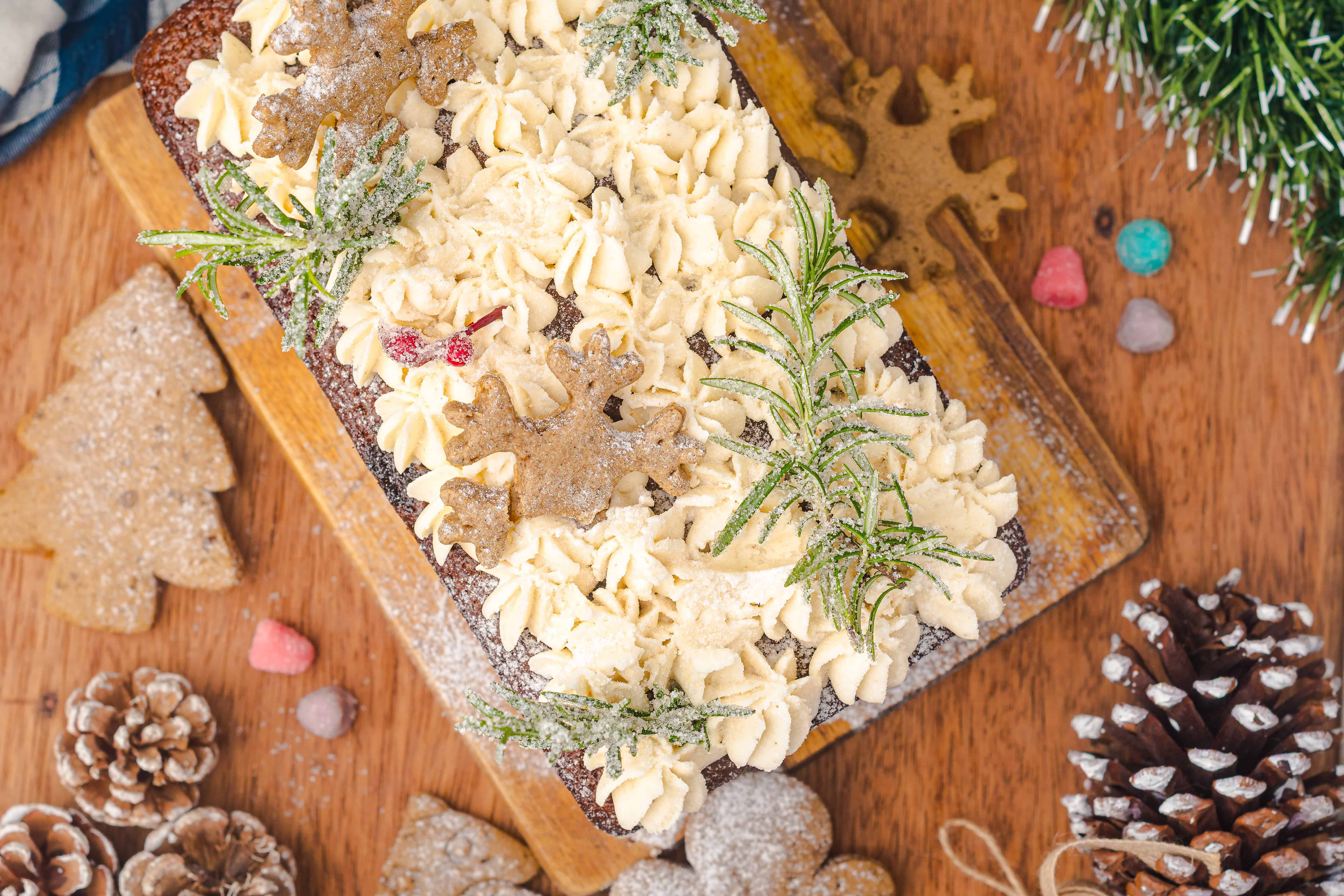 A festive Gingerbread Loaf cake with white frosting, gingerbread cookies, and sugared herbs served on a wooden board.