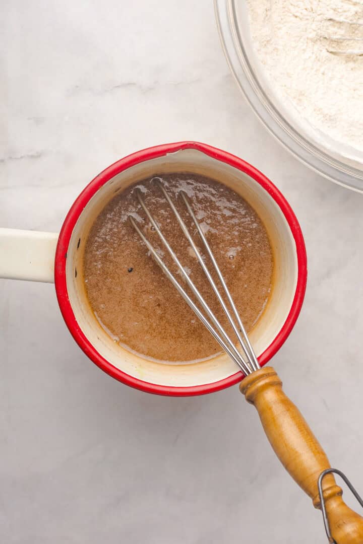 A whisk in a red saucepan with brown liquid, likely for a Gingerbread Loaf, sits next to a glass bowl of flour on a marble surface.