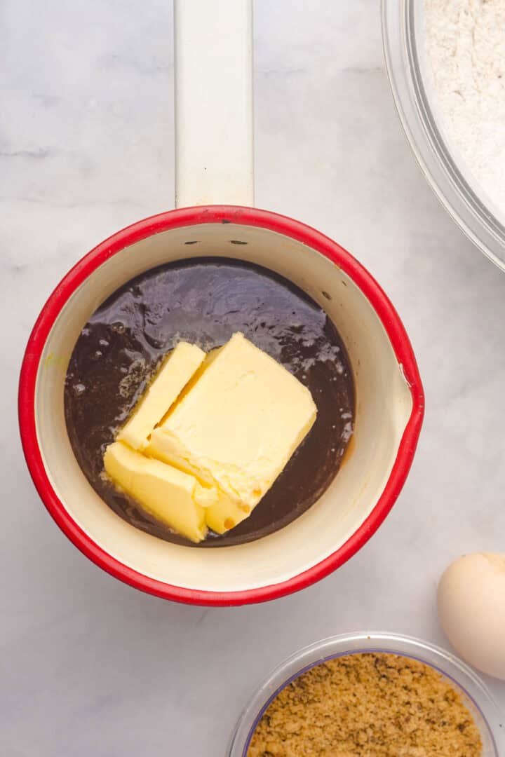 Butter melting in a small white saucepan with red rim, surrounded by baking ingredients for a gingerbread loaf on a marble surface.
