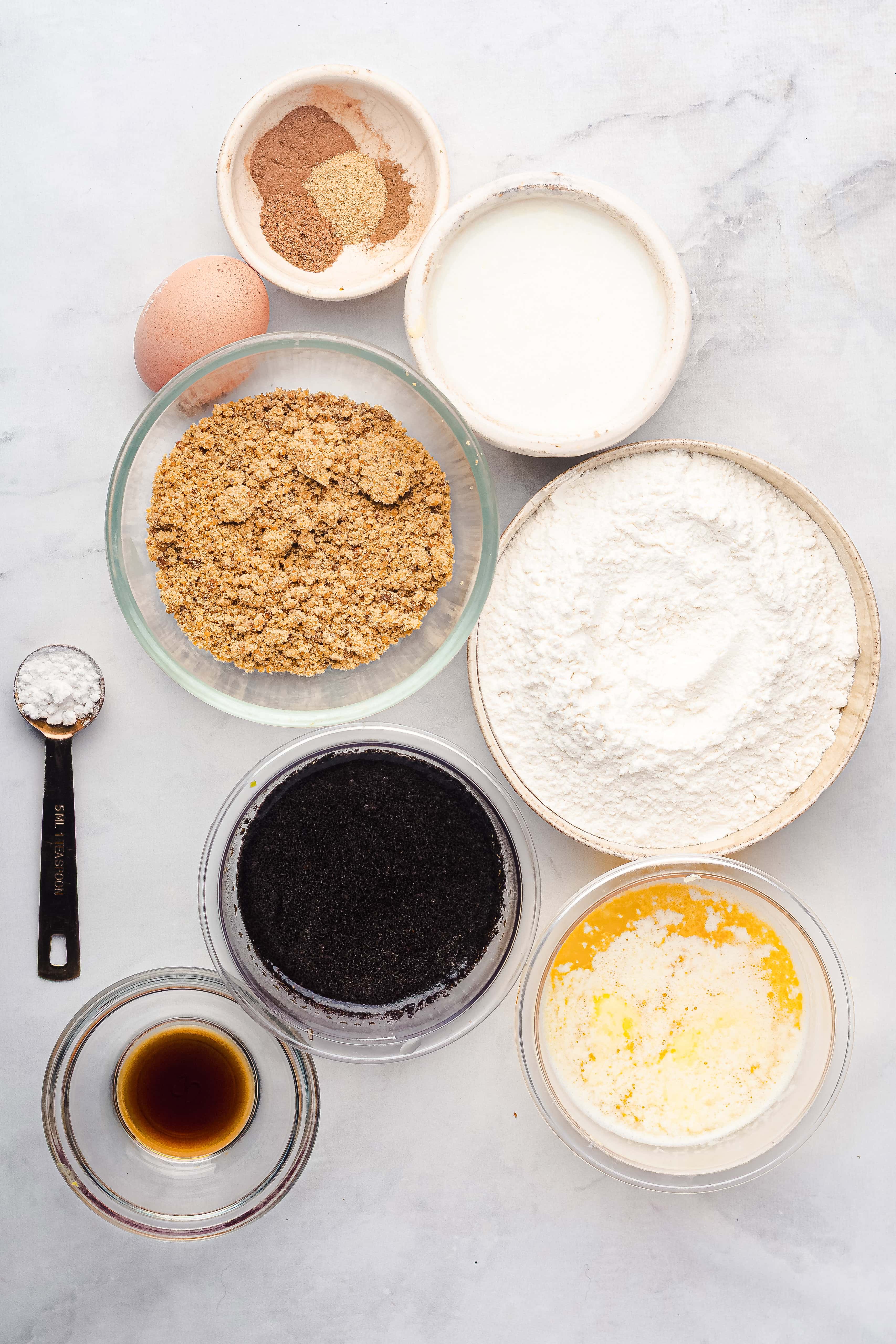 Baking ingredients for gingerbread muffins in bowls on a white surface: egg, milk, flour, poppy seeds, melted butter, spices, vanilla, and a teaspoon.