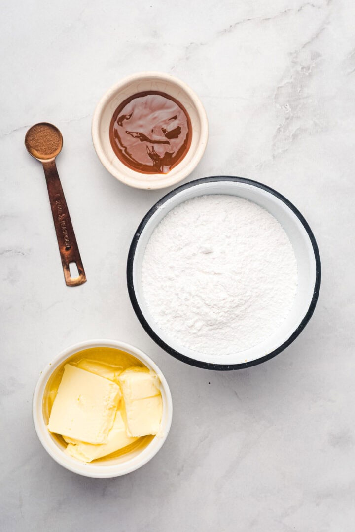 Four baking ingredients for gingerbread muffins: a bowl of powdered sugar, a bowl of butter, a bowl of honey, and a teaspoon of cinnamon.