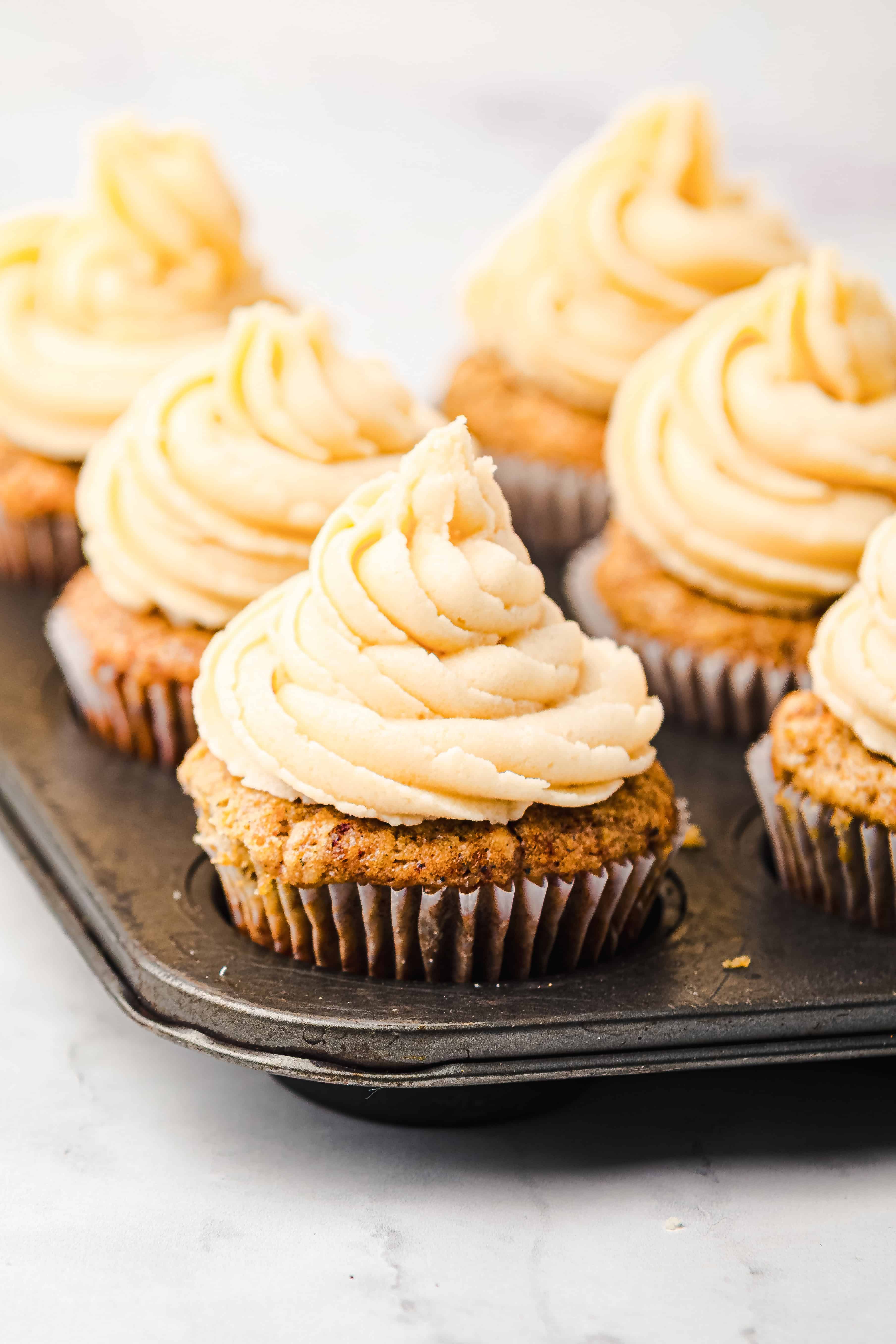 Gingerbread muffins with swirled frosting sit in a dark muffin tin on a light surface.