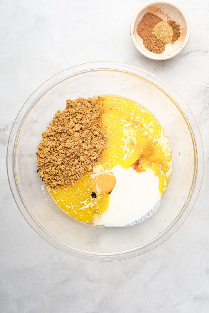 A glass bowl with brown sugar, egg, milk, and melted butter—perfect for gingerbread muffins; a small bowl of spices sits beside it.