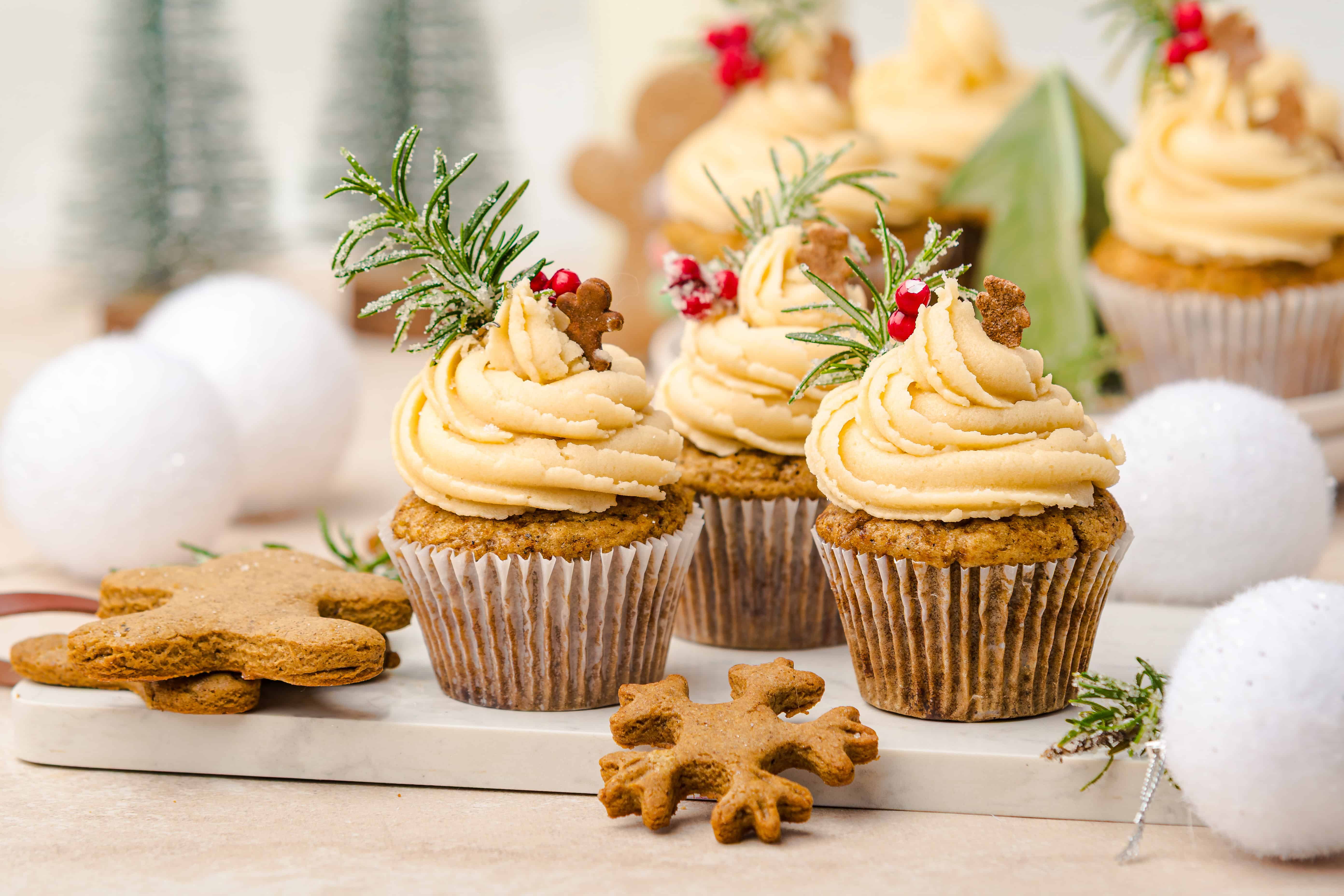Cupcakes with festive frosting, gingerbread muffins, cookies, and rosemary sprigs on a marble board, all surrounded by holiday decor.