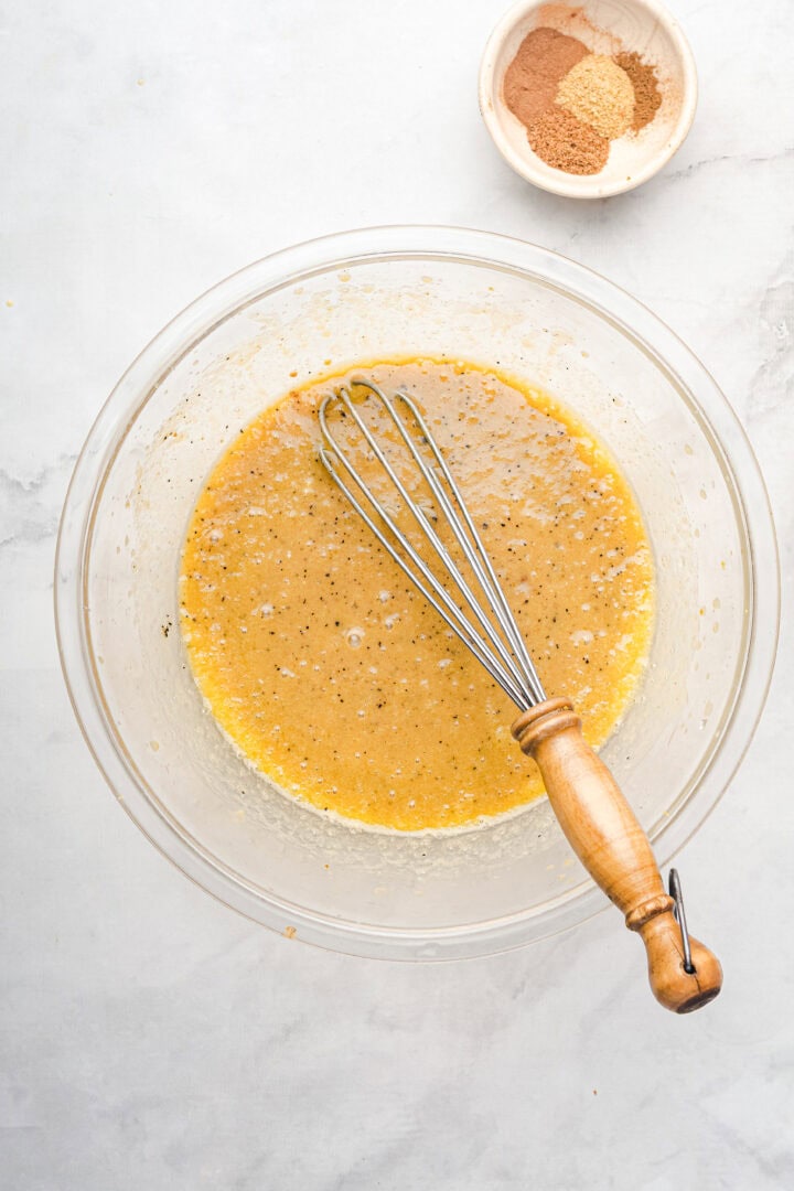 A whisk in a bowl of mixed batter for gingerbread muffins sits beside a small bowl of spices on a light surface.