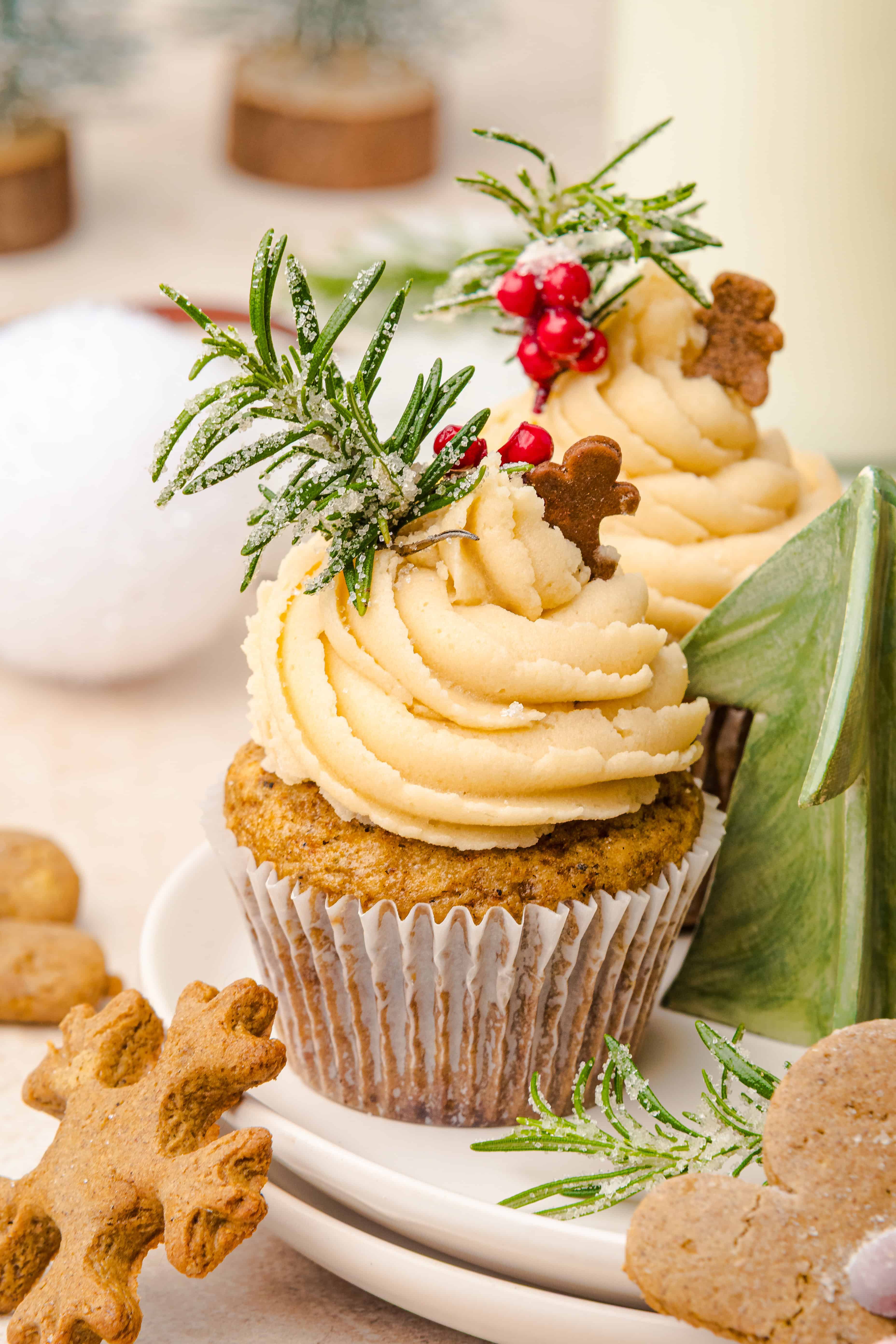 Cupcakes with swirled frosting, topped with rosemary, berries, and mini gingerbread muffins, arranged on a festive plate.