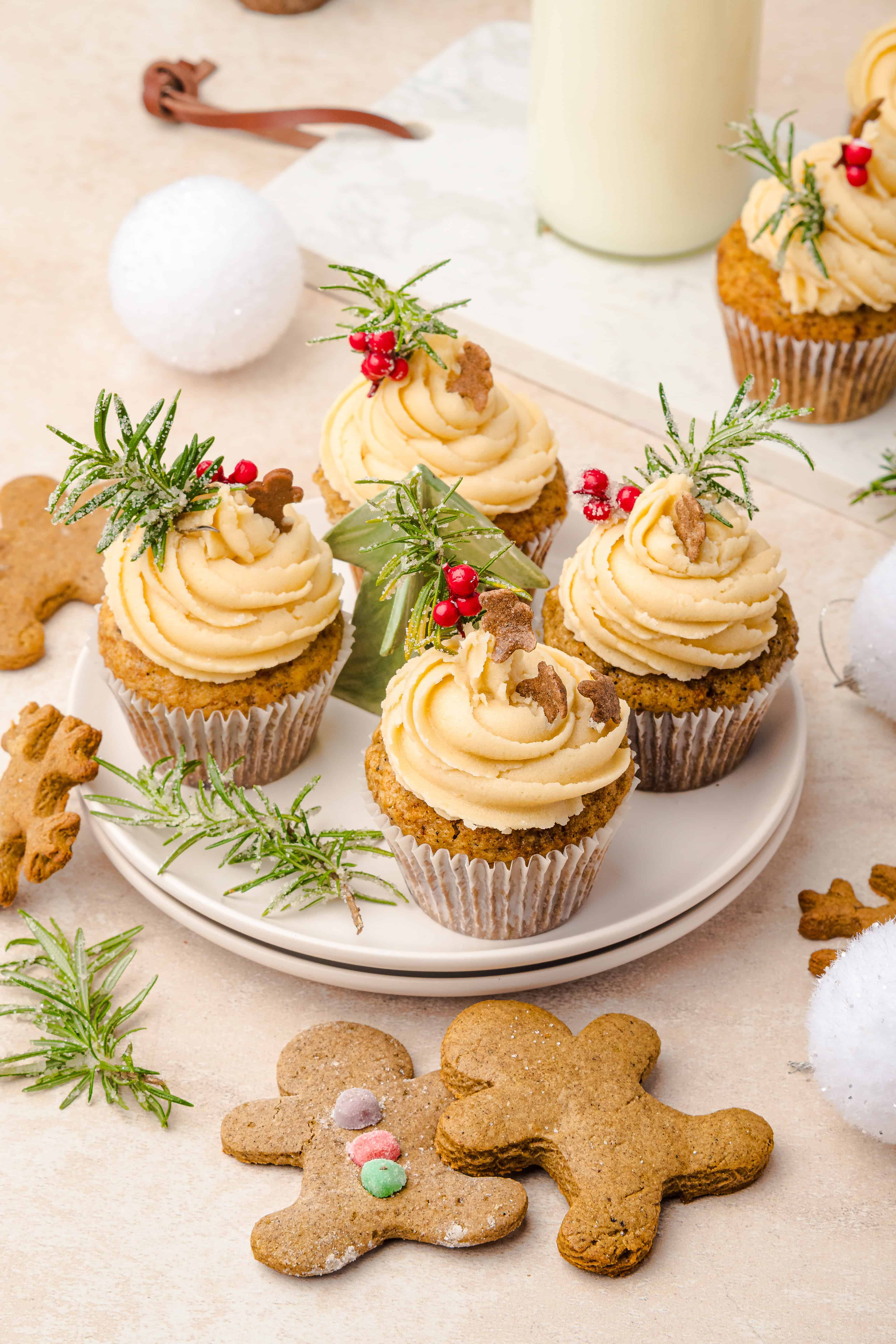 Cupcakes with creamy swirls, festive greenery, and red berries on a plate, surrounded by gingerbread muffins and cookies.