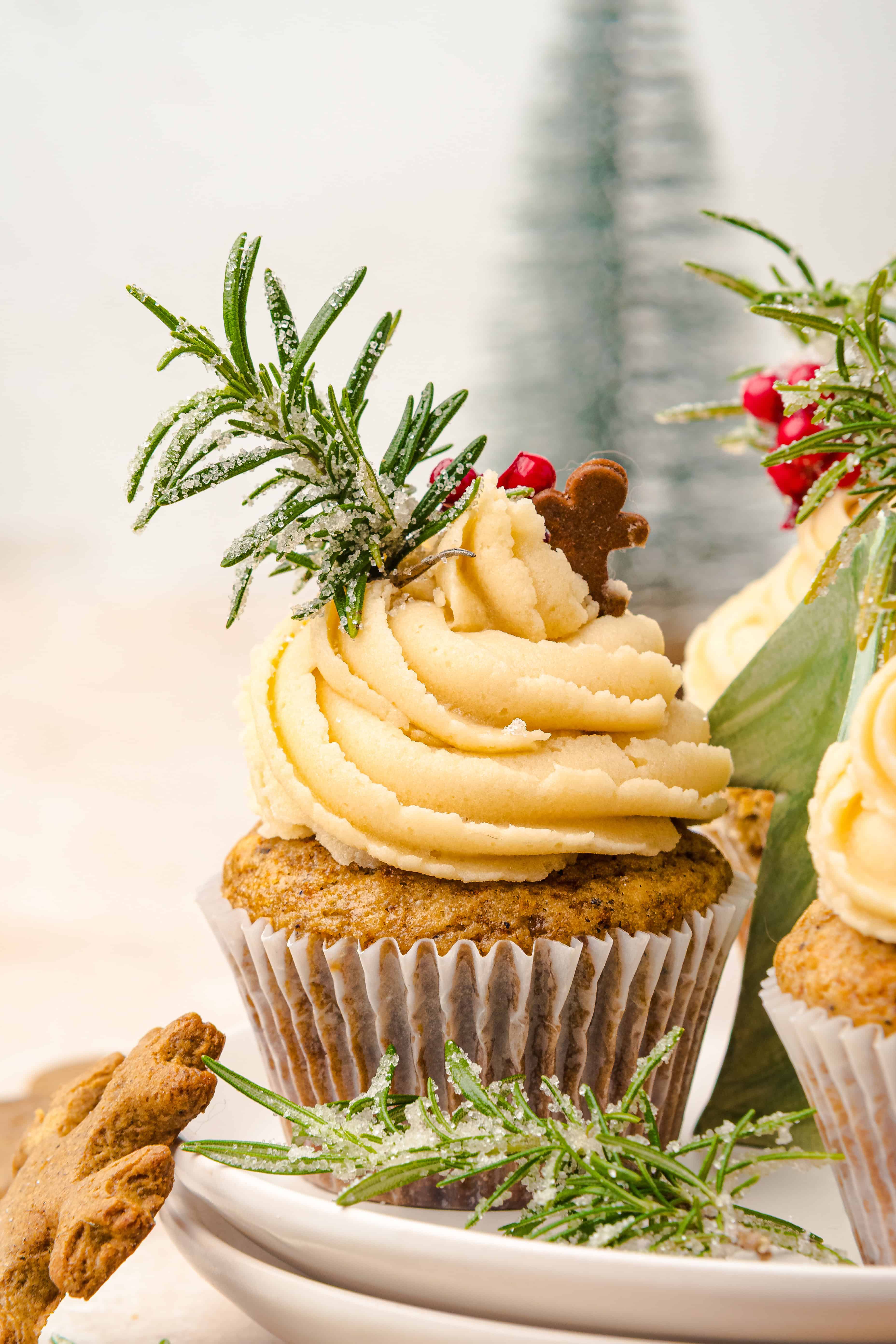 Cupcake with creamy frosting, rosemary, red berries, and a gingerbread cookie, reminiscent of festive gingerbread muffins, with a blurred tree in the background.