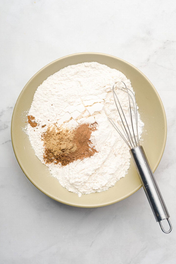 A bowl of flour and spices for gingerbread muffins sits with a metal whisk on a white surface.