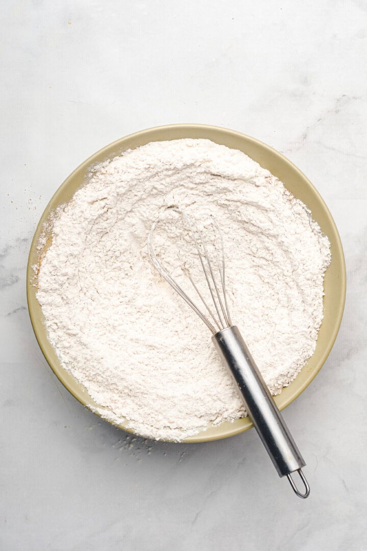 A metal whisk rests in a bowl filled with flour on a light-colored surface, ready for mixing up delicious gingerbread muffins.