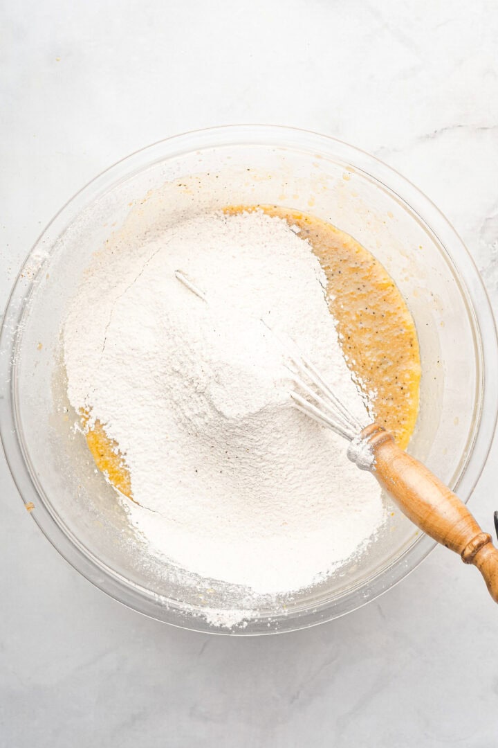 A whisk in a glass bowl with flour on top of a yellow batter, ready to be mixed for delicious gingerbread muffins.