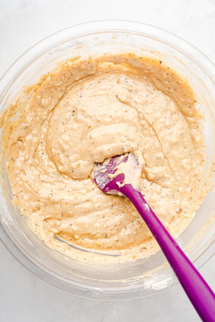 Thick, speckled gingerbread muffin batter being mixed in a glass bowl with a purple spatula on a white surface.